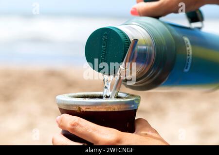 Femme servant l'eau chaude d'un thermos Stanley dans un Mate avec yerba. Boire Mate sur la plage. Mar Azul, Buenos Aires, Argentine - 25 janvier 2024. Banque D'Images