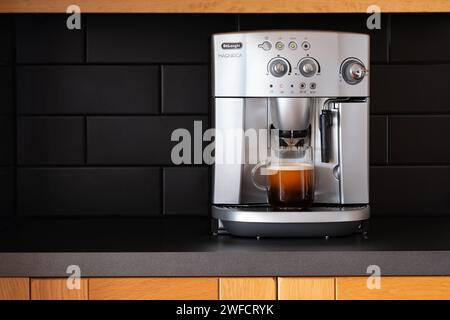 Une machine à café domestique Delonghi Magnifica UK à grain à tasse sur un plan de travail de cuisine. Un café fraîchement préparé dans une tasse en verre se trouve sur le plateau de la machine Banque D'Images