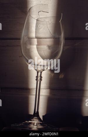 Verre à vin cassé dans la lumière du soleil sur fond en bois. Verre à vin endommagé avec bords tranchants. Verre fissuré dans les ombres. Nature morte créative. Objet fragile Banque D'Images