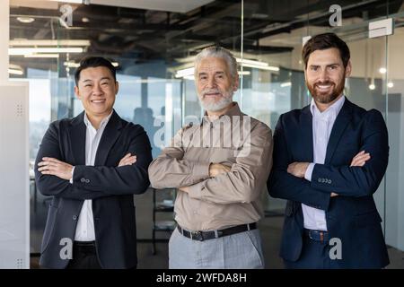 Trois hommes professionnels confiants debout ensemble les bras croisés dans un environnement de bureau contemporain, rayonnant travail d'équipe et leadership. Banque D'Images