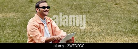 heureux aveugle indien en veste assis sur l'herbe avec des lunettes et lisant le code braille, bannière Banque D'Images