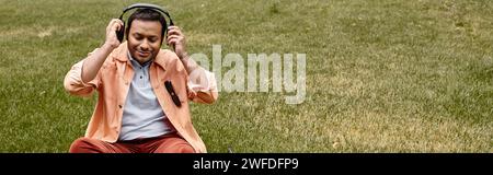 homme aveugle indien joyeux dans la veste orange assis sur l'herbe avec des écouteurs et des lunettes, bannière Banque D'Images