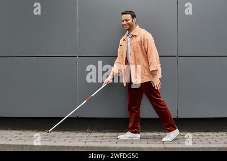 homme aveugle indien joyeux dans la veste orange avec aide bâton marchant avec mur gris en toile de fond Banque D'Images