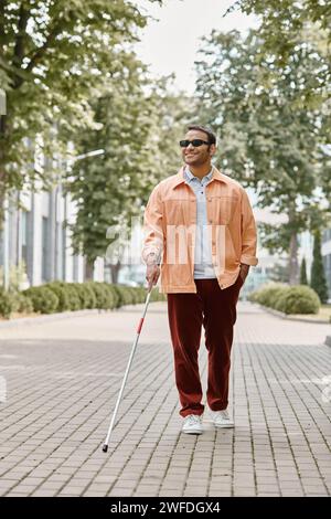 homme aveugle indien joyeux avec des lunettes et bâton de marche dans la veste vive orange marchant dans le parc Banque D'Images