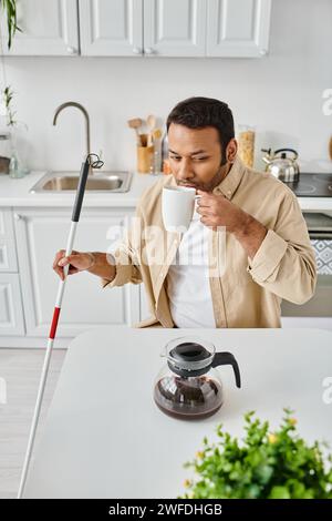 bel homme indien avec déficience visuelle assis et boire un café savoureux alors que sur la cuisine Banque D'Images