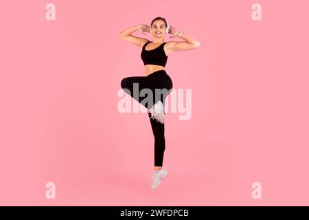Jeune femme active portant des écouteurs sautant sur fond de studio rose Banque D'Images