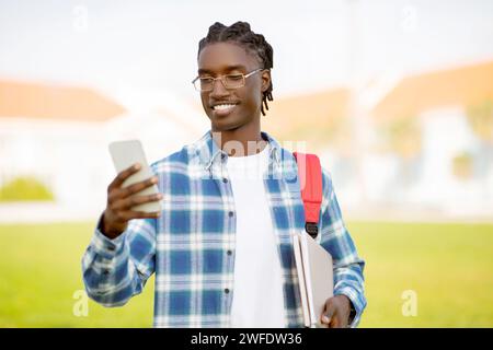 Étudiant noir heureux tient smartphone, chats et études en ligne à l'extérieur Banque D'Images