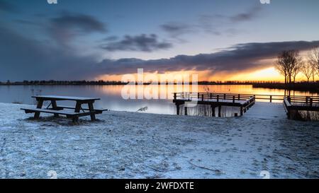 Jetée et table de pique-nique en bois au bord du lac Dirkshorn dans un cadre hivernal. La neige pendant la nuit crée un paysage blanc au lever du soleil. Une averse de neige à l'horizon. Banque D'Images
