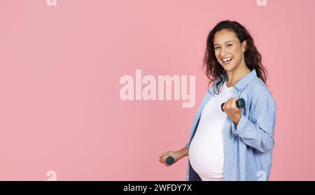 Souriant femme brune enceinte s'exerçant avec des haltères sur fond rose Banque D'Images