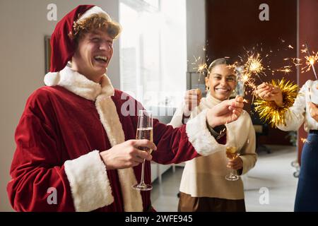 Jeune homme d'affaires riant en costume de Père Noël tenant l'étincelle brûlante et flûte de champagne tout en appréciant la célébration de Noël Banque D'Images