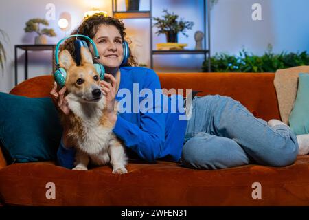 Heureuse jeune femme écoutant de la musique à travers des écouteurs sans fil couchés sur le canapé avec chien corgi. Fille féminine joyeuse avec les cheveux bouclés dansant sur la musique dans le salon à la maison. Musique de danse Banque D'Images