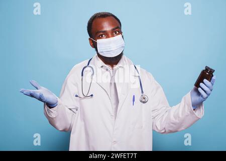 Chimiste confus tenant un contenant antidouleur tout en portant un masque facial, un stéthoscope et des gants en latex. Médecin perplexe, saisissant un flacon de pilule d'ordonnance et vêtu d'une blouse de laboratoire. Banque D'Images