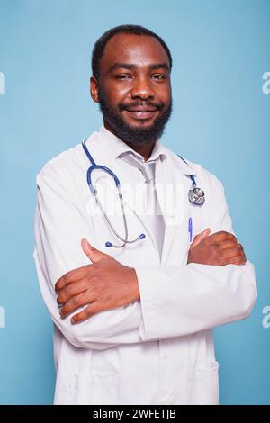 Médecin souriant d'ethnie afro-américaine ayant les bras croisés est positionné devant un fond bleu isolé. Spécialiste de la santé masculine avec stéthoscope posant pour la caméra en studio. Banque D'Images