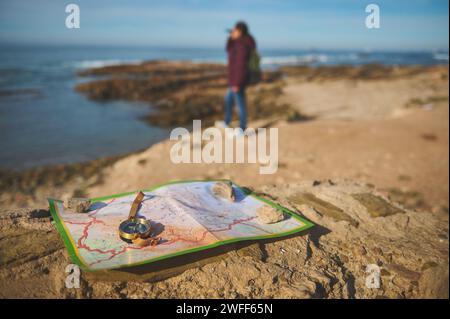 Détails sur une boussole et carte sur un rocher contre touriste féminine floue regardant dans la distance à travers des jumelles, debout sur le sommet d'une falaise, annonce Banque D'Images