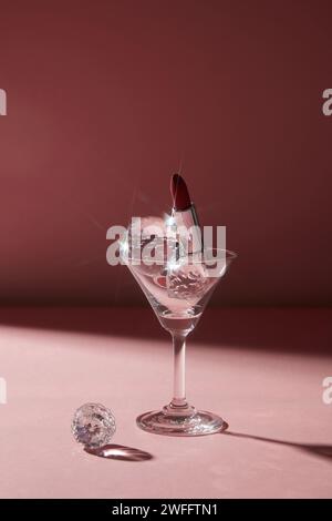 Un verre à cocktail transparent contenant une sphère de diamant et un élégant rouge à lèvres rouge dans un emballage argenté. Fond sombre pour le concept de nuit de dame Banque D'Images