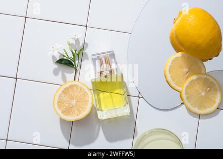 Sur un fond de carreaux de mosaïque blanche, un flacon transparent au parfum présenté avec une branche de fleur, un miroir et une boîte de Pétri de liquide jaune. Citron (C Banque D'Images