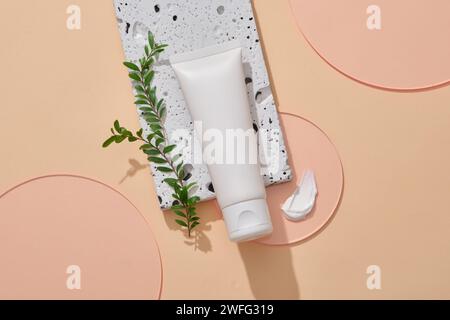 Un tube cosmétique à étiquette vide posé sur un podium en pierre, décoré avec des feuilles, une texture crème et quelques feuilles acryliques roses. Maquette de produit de marque beauté Banque D'Images