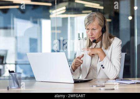 Une femme d'affaires senior sérieuse et ciblée mène une réunion vidéo en ligne avec des clients et des partenaires. Assis dans le bureau au bureau dans un casque et discuter du travail. Banque D'Images