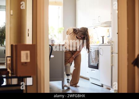 Femme mature avec handicap vérifiant la jambe prothétique tout en se tenant dans la cuisine à la maison Banque D'Images