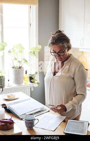 Femme mature photographiant les factures financières tout en se tenant dans la cuisine à la maison Banque D'Images