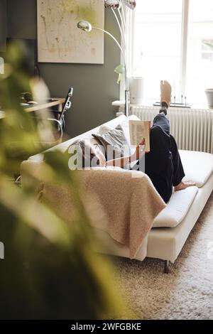 Femme mature lisant le livre tout en se couchant sur le canapé à la maison Banque D'Images