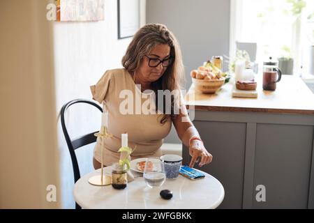 Femme mature avec handicap défilant téléphone intelligent tout en étant assis à la table prenant le petit déjeuner à la maison Banque D'Images