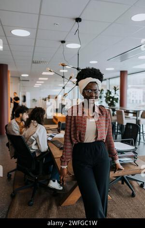 Portrait de jeune femme d'affaires souriante appuyée sur le bureau contre des collègues discutant de stratégie dans le bureau Banque D'Images