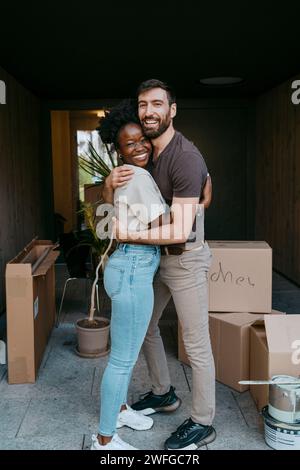 Portrait vue de côté d'un couple multiracial heureux s'embrassant tout en se tenant debout à l'entrée du bâtiment Banque D'Images