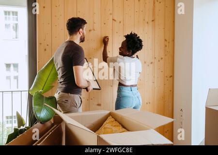 Heureux femme martelant clou avec petit ami tenant le cadre près du mur en bois à la nouvelle maison Banque D'Images