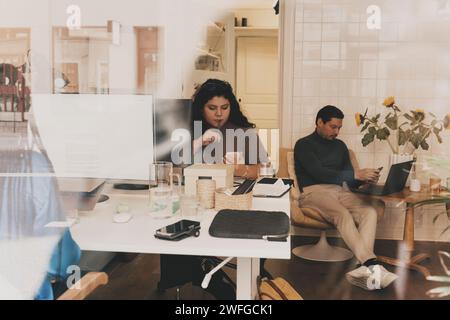 Femme d'affaires réfléchie utilisant le téléphone intelligent tout en étant assis au bureau vu à travers la fenêtre en verre Banque D'Images