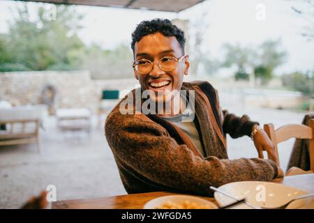 Heureux jeune homme portant des lunettes assis à la table à manger dans le patio Banque D'Images