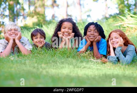 Nature, sourire et portrait d'enfants sur l'herbe dans le parc extérieur, le champ ou le jardin ensemble. Heureux, diversité et groupe de jeunes enfants excités se relaxant Banque D'Images