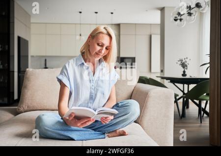 Femme assise confortablement sur le canapé, profondément engagée dans la lecture du livre. Espace de vie confortable avec une esthétique moderne sur fond. Personne jouissant d'un moment calme de solitude, immergé dans la lecture. Banque D'Images