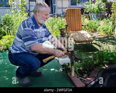 Un homme aux cheveux gris et un samovar. Un homme coupe des copeaux de bois pour un samovar dans le jardin. Thé dans le jardin au samovar. Banque D'Images