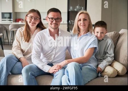 Grande famille se refroidissant ensemble sur un canapé crème. Portrait de famille mignon de quatre membres regardant l'appareil photo. Heureux moments de personnes proches dans le salon moderne. Banque D'Images