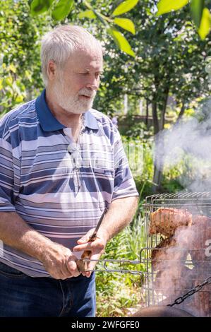 Un homme aux cheveux gris fait griller de la viande sur un gril près du brasero. La viande est cuite sur un barbecue maison dans la nature. Banque D'Images