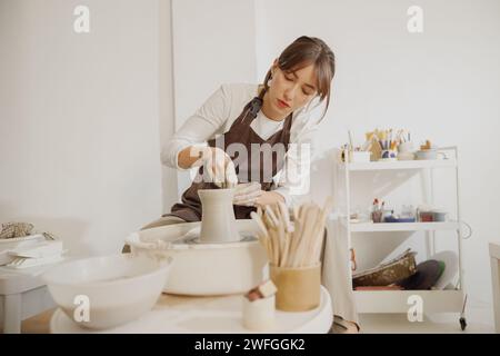 Artisan féminin concentré en tablier assis sur un banc avec roue de poterie et faisant pot d'argile Banque D'Images