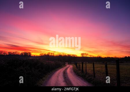 Une piste de terre sinueuse passant à travers les champs et menant vers une forêt et un lever de soleil orange et violet dramatique dans la région de Dordogne en France Banque D'Images