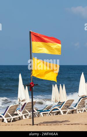 Des drapeaux rouges et jaunes flottent sur une plage avec des chaises longues pliées et des parasols fermés en arrière-plan signalant des conditions de baignade à risque moyen Banque D'Images