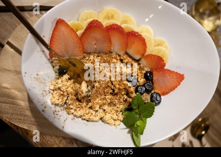 Bouillie chaude de flocons d'avoine ornée de fraises mûres, de tranches de banane, de myrtilles et d'un généreux filet de miel doré. Banque D'Images