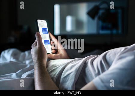 Téléphone dans le lit. Chat par SMS sur l'écran du smartphone la nuit. Homme dans la chambre à coucher sombre de la maison ou la chambre d'hôtel. Rencontres en ligne, escroquerie mobile sms ou secret. Banque D'Images