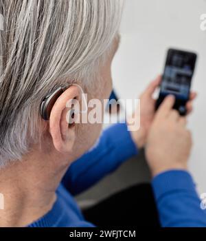 Homme senior malentendant qui règle les paramètres de son aide auditive BTE via un smartphone. Prothèses auditives avec technologies innovantes en audiologie Banque D'Images