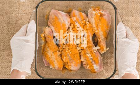 Les mains du chef portant des gants tenant un plat de cuisson en verre avec des pilons de poulet marinés dedans. Recette de pilons de poulet cuits au four, préparation p Banque D'Images