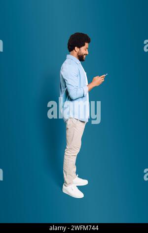 Un jeune homme de profil, profondément engagé avec son téléphone, illustrant la nature omniprésente des appareils mobiles dans la vie moderne. Photo en longueur de l'homme brésilien à l'aide d'un smartphone isolé sur bleu Banque D'Images