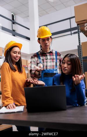 Équipe de travailleurs de magasin asiatique analysant la liste de contrôle d'expédition ensemble et discutant des opérations d'expédition de commande. Employés souriants de l'entrepôt recherchant des informations sur les produits sur ordinateur portable Banque D'Images