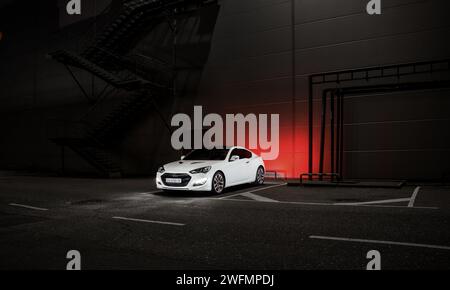 Blanc Hyundai Genesis coupé garé près du mur gris et des escaliers d'évacuation de secours. Vue de trois quarts de nuit de la photo du coupé sportif coréen dans la zone industrielle. Banque D'Images