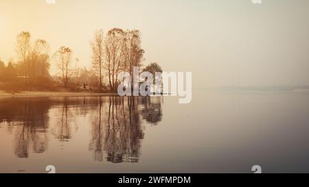 Arbres sur la rive de la rivière au lever du soleil. Reflet symétrique de quelques arbres dans l'eau par un matin brumeux. Banque D'Images