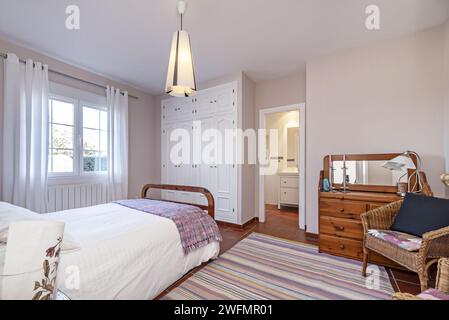 Une chambre avec une couette blanche avec des coussins à côté de la fenêtre et une armoire encastrée en bois blanc à côté de la salle de bains Banque D'Images