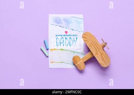 Carte pour la fête des pères avec craies et avion en bois sur fond lilas Banque D'Images