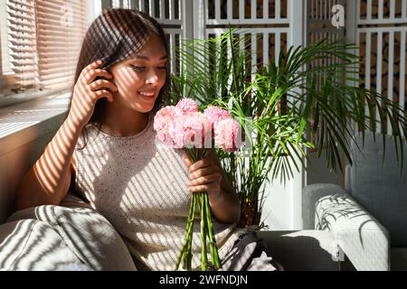 Belle jeune femme asiatique avec bouquet de dahlias roses assis dans le salon près de la fenêtre Banque D'Images